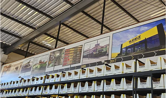 Painel em lona com a linha do tempo da BBZ Materiais Elétricos, de 2009 a 2017, acima das prateleiras do galpão